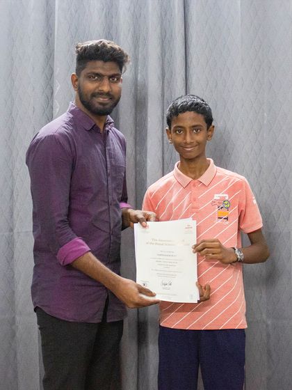 A student proudly holds his ABRSM exam certificate next to his teacher.