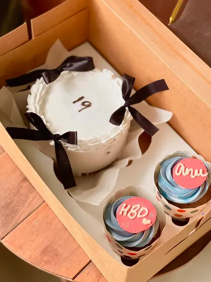 A chic and minimalist 19th birthday combo. The white bento cake is decorated with delicate black bows, and the two cupcakes feature a lovely blue frosting with personalized pink toppers.