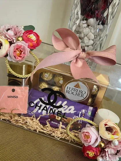 A close-up of the Valentine's hamper, showcasing the mix of high-end chocolates, Miniso earrings, and a candle. The faux Chanel logo adds a fun, fashionable touch to the arrangement.