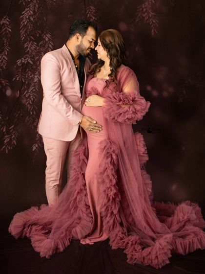 A classic couple pose, standing together, both cradling the baby bump. The coordination in their outfits adds to the elegance of this studio portrait.