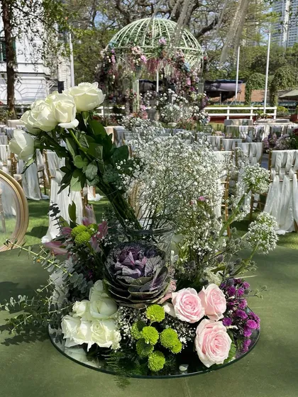 A beautiful floral centerpiece for a garden wedding, featuring white roses, baby's breath, and soft pink and purple blooms. The mirrored base reflects the surrounding greenery, adding depth to the table decor.
