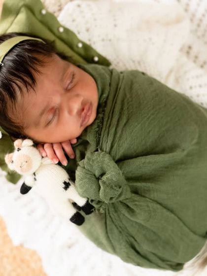 A sleeping newborn in an olive green wrap, cuddling a tiny toy sheep in a rustic basket.