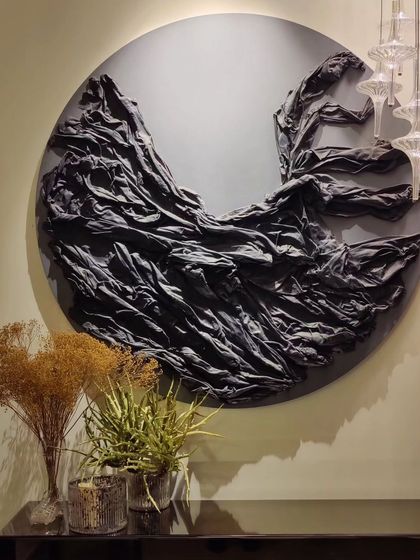 'LUNA' is a circular wall sculpture that explores texture and form. Created using plaster-dipped fabric, the piece mimics the dramatic, flowing movement of cloth frozen in time, set against a smooth, dark background.