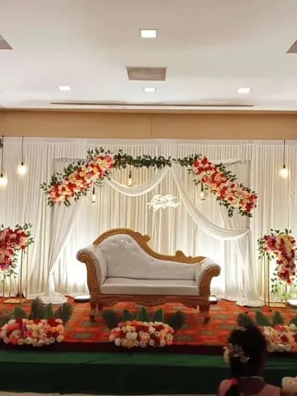 A grand stage decoration for a formal engagement or reception in a hall. The design features a wide white curtain backdrop, an arch of red and white flowers, and a classic white chaise lounge for elegant seating.