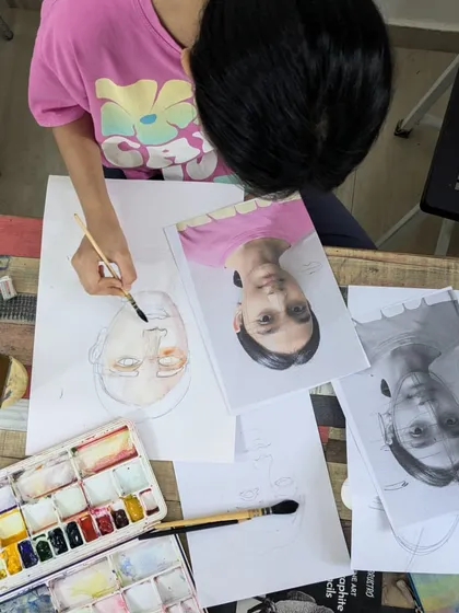 A student working on a watercolor portrait. This overhead view shows the process of layering skin tones and using reference photos for accuracy.