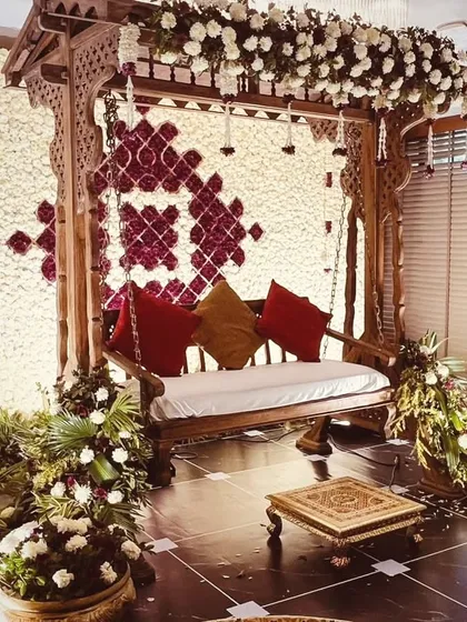 A classic and ornate wooden jhoola (swing) set against a dense wall of white and burgundy flowers. This traditional seating arrangement is perfect for Mehendi or Haldi ceremonies, offering a touch of royal elegance.