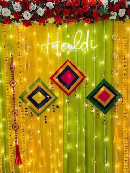 A bright and colorful Haldi backdrop with a neon sign. The decor features yellow and lime green drapes, marigold garlands, and multi-colored hangings, perfect for a fun and modern celebration.