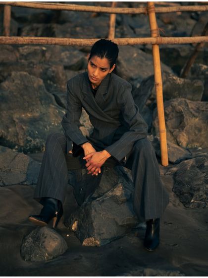 A full-length shot of the model seated on beach rocks. This pose feels both powerful and relaxed, showcasing the versatility of the styling in a natural setting.