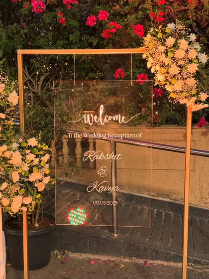 A simple and elegant acrylic welcome sign for Rakshit and Kavya's wedding reception, framed by soft flowers and greenery.