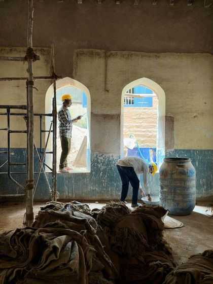 The classroom interior during restoration, with its arched windows and high ceilings. Scaffolding fills the space as workers prepare the walls for a new coat of lime plaster, bringing the room back to its original proportions and light.