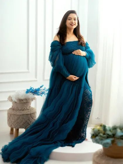 A full-length portrait showcasing an elegant, deep teal gown. The off-shoulder design and flowing train create a look of modern sophistication.