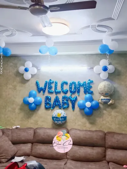 A classic 'Welcome Baby' boy decoration for a living room. The setup includes blue and white balloons, flower-shaped balloon clusters, and 'WELCOME BABY' foil letters.