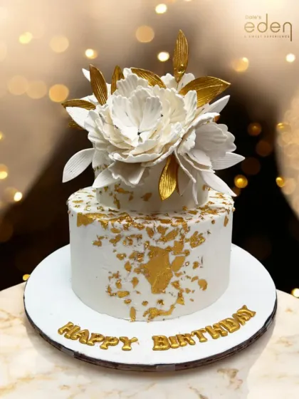 A masterpiece of elegance. This white birthday cake is topped with a large, handcrafted white sugar flower with golden-edged petals, making it a sophisticated and artistic centerpiece.