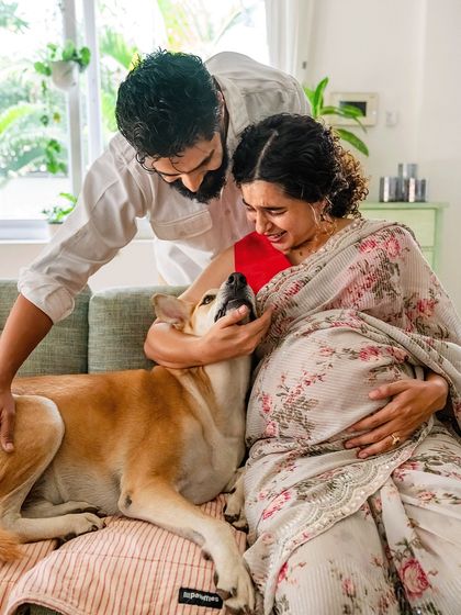 A moment of pure, unscripted love. An expectant mother laughs as her husband and their dog share a sweet, affectionate interaction during their at-home maternity session.