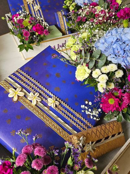 A close-up of a beautifully wrapped gift, showcasing the detailed ribbon work with shell flowers and gota trim. The surrounding fresh flowers in shades of blue, pink, and purple complete the luxurious look.