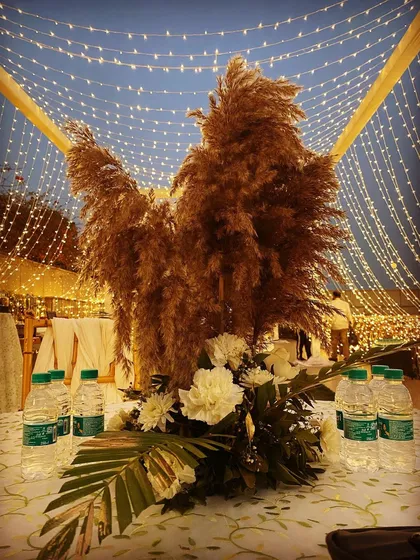 A beautiful centerpiece for a boho-themed table. A large plume of pampas grass is paired with white flowers, set against a backdrop of fairy lights for a magical evening look.