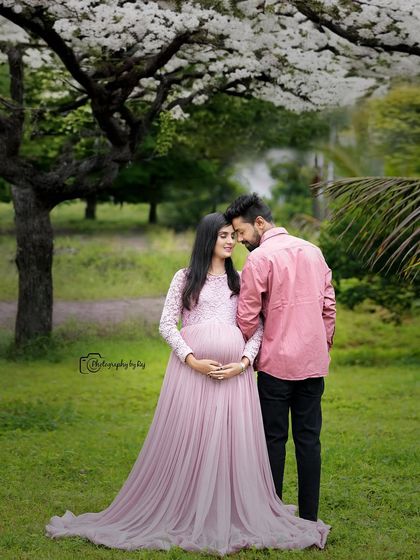 A romantic couple's portrait under a tree with white blossoms. The mother-to-be's light pink lace gown adds to the soft, dreamy feel of the image.
