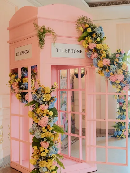A whimsical pink telephone booth covered in a cascade of colorful flowers, serving as a fun and highly instagrammable photo op.