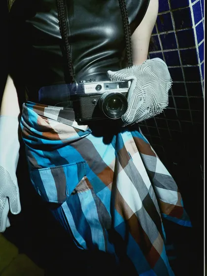 A detail shot focusing on the vintage camera as a prop, tying into the 'Peek-a-boo' theme. The textures of the plaid skirt, leather top, and gridded gloves are prominent.