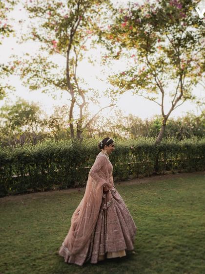 The bride walks through a garden at The Ananta Udaipur, her pink lehenga glowing in the soft light. This wide shot captures a moment of quiet grace and beauty.