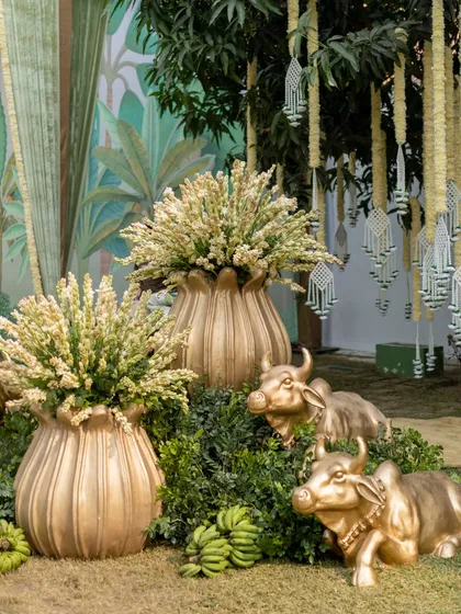 A close-up of the decor, featuring golden cow statues nestled in greenery, bunches of green bananas, and brass urns filled with tuberose, all elements of a traditional and prosperous celebration.