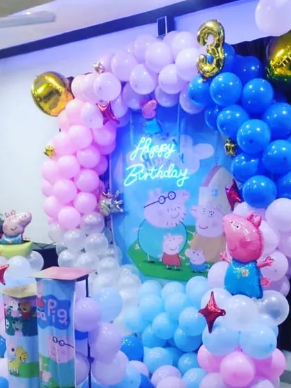 A cheerful Peppa Pig themed birthday party. The decoration features character cutouts against a themed backdrop, surrounded by a colorful balloon arch in shades of pink and blue.