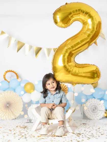 A series of portraits celebrating a second birthday. The toddler poses with a giant gold '2' balloon, surrounded by blue and white balloons and confetti for a festive look.