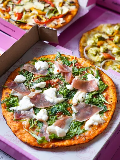 A delivery box open to reveal a delicious prosciutto and arugula pizza, with other pizzas in the background. This shot is perfect for a delivery or takeaway menu, showcasing variety.