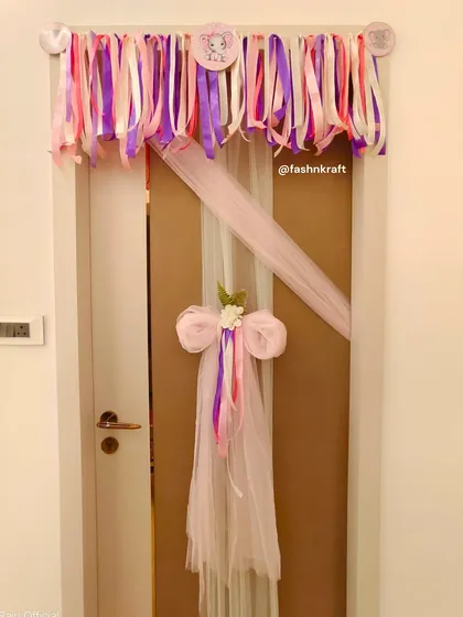 A playful and pretty decoration for a baby girl's room door. A colorful ribbon garland hangs above, while a large tulle bow with trailing ribbons adds a touch of whimsy and charm.