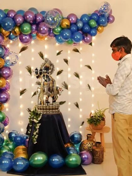 For a home puja or Janmashtami celebration, I can create a beautiful and sacred space. This setup features a balloon arch in peacock-inspired colors, a backdrop of fairy lights and feathers, and a decorated pedestal for the Krishna idol.