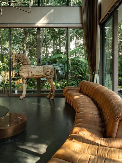 A double-height living room with expansive glass walls that look out onto a dense garden and water body. The interior feels vast and open, directly connected to the tranquil landscape outside.