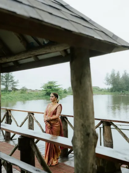 A serene portrait of a South Indian bride in her beautiful silk saree, standing on a wooden pier by the water. The natural setting complements her timeless elegance.
