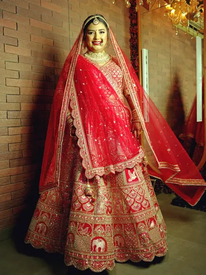 The classic full-length bridal portrait. This shot showcases the bride's entire stunning red lehenga, with its intricate elephant motifs, against a simple brick wall that makes the outfit's colors and details stand out.