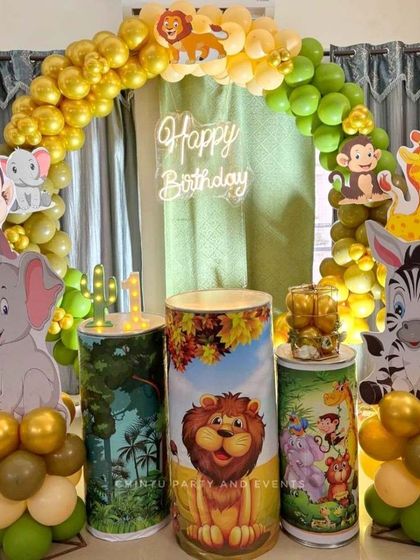 This jungle-themed birthday setup features cute animal cutouts like an elephant and a monkey, themed pedestals for the cake, and a balloon arch in green and gold. A 'Happy Birthday' neon sign completes the look.
