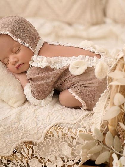 A newborn in a beautiful beige lace outfit sleeps on a miniature rattan bed, styled with dried white flowers for a delicate and dreamy look.