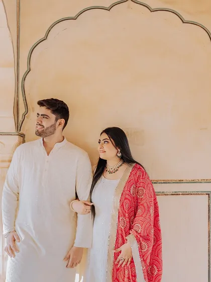 A classic portrait of a couple in elegant white attire, with the bride's vibrant red dupatta adding a beautiful pop of color.