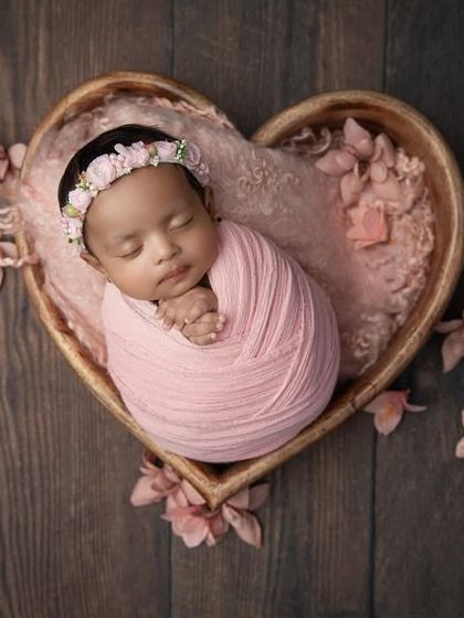 A sweet baby girl swaddled in soft pink, nestled in a heart shaped bowl on a dark wood background. The delicate flowers add a romantic, gentle touch.