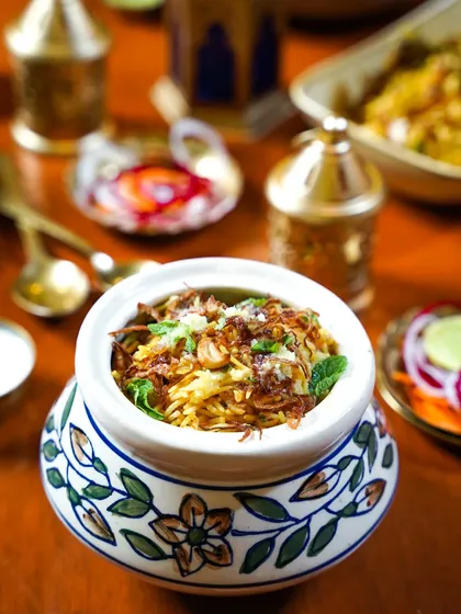 A beautifully styled pot of biryani, garnished with fried onions and mint. The traditional ceramic handi and warm tones evoke a sense of authentic, slow-cooked flavor.