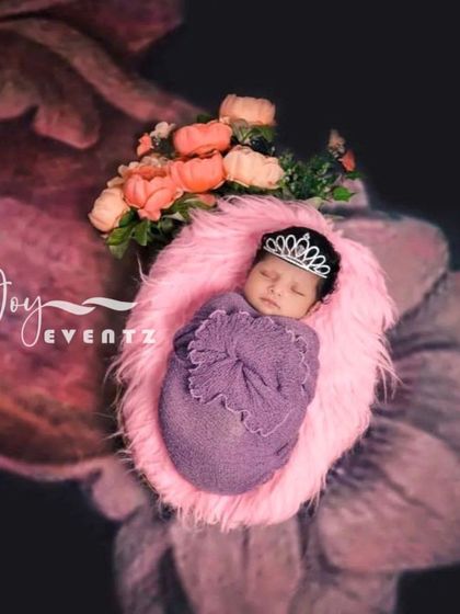 A princess-themed newborn portrait where the baby, wearing a tiny tiara, is swaddled in purple and rests on a soft pink fur cushion. The floral background adds to the royal and delicate feel of the photograph.