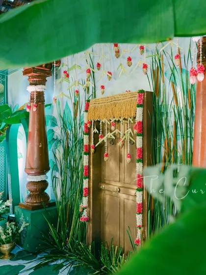 Your Muhurtham, our canvas. This artistic shot, framed by banana leaves, shows our custom-designed backdrop featuring a painted reed motif and a traditional door, blending art with celebration.