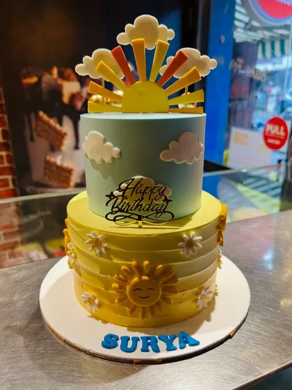 A bright and happy two-tier "sunshine" themed cake for Surya, with a smiling sun and fluffy clouds.