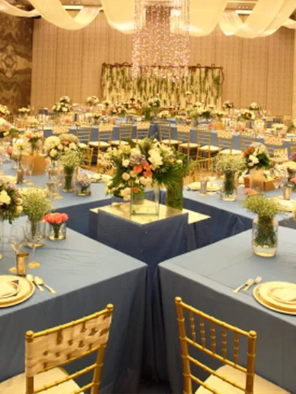 A reception hall transformed with stunning blue linens and gold chiavari chairs. The square table formation encourages conversation, while the grand chandelier adds a touch of timeless elegance.
