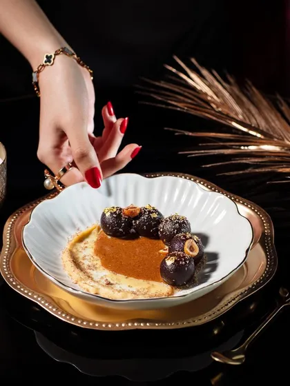 An elegant dessert shot of Kala Gulab Jamun for Club Jolies. The composition is luxurious, with gold-rimmed plates and soft candlelight, perfectly matching the fine dining and fusion concept of the dish.