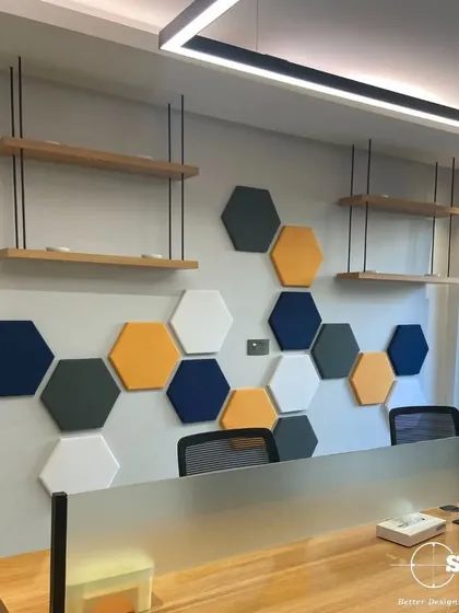 A collaborative workspace featuring a honeycomb-patterned acoustic wall panel. The design uses a playful mix of colors and suspended wooden shelves to create a dynamic and creative environment.