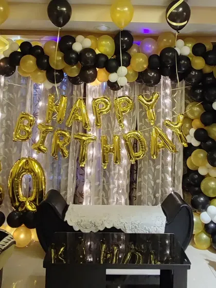A 50th birthday celebration with a classic black and gold theme. The decoration includes a full balloon garland, large '50' foil numbers, and 'Happy Birthday' foil letters against a simple curtain backdrop.