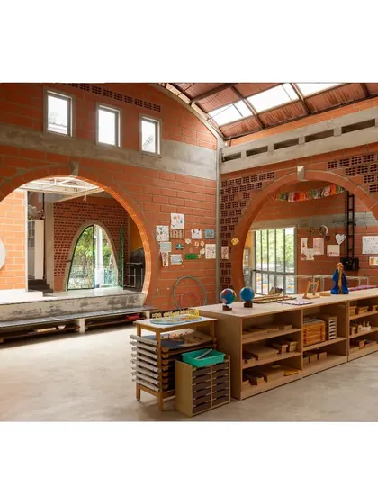 The interior learning space is defined by its honest materials and grand scale. Soaring arches made of hollow clay blocks create visual pathways, while ample shelving and open tables provide a functional environment for Montessori-style education.