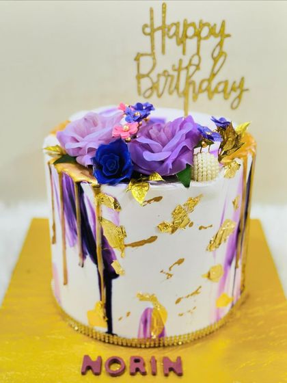 A striking white cake with bold purple and gold brushstrokes, topped with a beautiful arrangement of purple and blue sugar flowers.