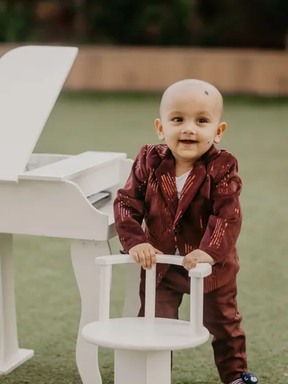 A charming photo of a baby boy in a velvet suit standing next to a mini white piano, smiling for the camera.