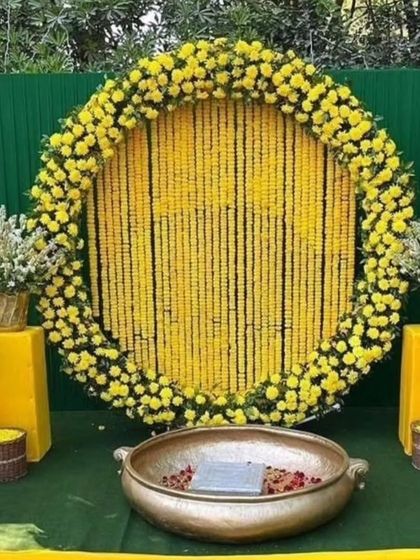 A classic Haldi backdrop for outdoor functions. This setup features a large circular frame filled with strings of yellow marigolds, a green panel background, and a traditional brass urli in the center for the ceremony.