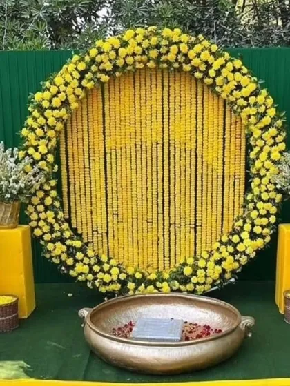 A classic Haldi backdrop for outdoor functions. This setup features a large circular frame filled with strings of yellow marigolds, a green panel background, and a traditional brass urli in the center for the ceremony.